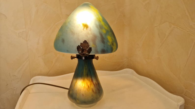 May include: A mushroom-shaped table lamp with a glass shade. The shade is a mix of blue, yellow, and green, with a leaf-shaped accent. The base is a dark teal color. The lamp is on a white surface.