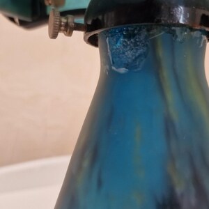 May include: Close-up of a teal and blue glass object with a flared base. The glass has a textured, mottled appearance. The object is topped with a black metal fixture with adjustable screws.