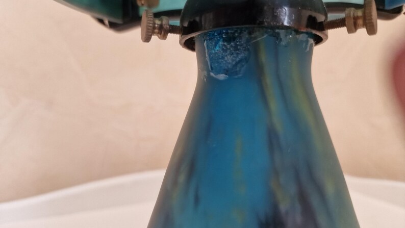 May include: Close-up of a teal and blue glass object with a flared base. The glass has a textured, mottled appearance. The object is topped with a black metal fixture with adjustable screws.