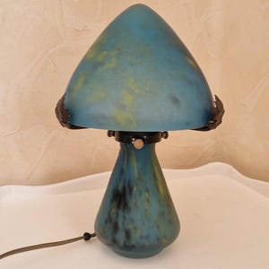 May include: A blue mushroom-shaped table lamp with a conical shade and a rounded base. The lamp has a mottled blue and yellow color scheme, with dark accents. The lamp is on a white surface.