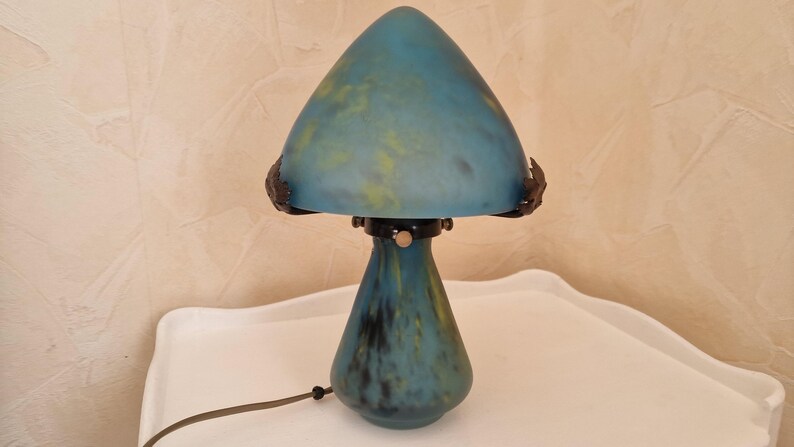 May include: A blue mushroom-shaped table lamp with a conical shade and a rounded base. The lamp has a mottled blue and yellow color scheme, with dark accents. The lamp is on a white surface.