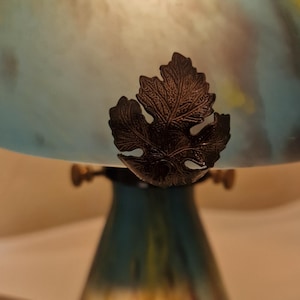 May include: A decorative table lamp with a blue and yellow glass shade. The lamp features a bronze-colored leaf accent and a teal-colored base. The shade has a bear silhouette. The lamp is designed to provide soft, ambient lighting.