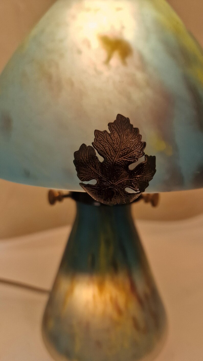 May include: A decorative table lamp with a blue and yellow glass shade. The lamp features a bronze-colored leaf accent and a teal-colored base. The shade has a bear silhouette. The lamp is designed to provide soft, ambient lighting.