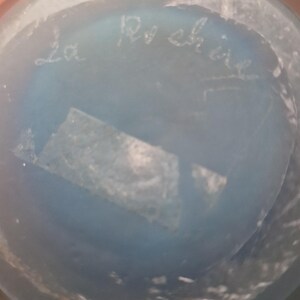 May include: A close-up of a light blue glass object with the handwritten text "La Roch&egrave;re". The object has a slightly textured surface and a small, rectangular piece of tape is visible.
