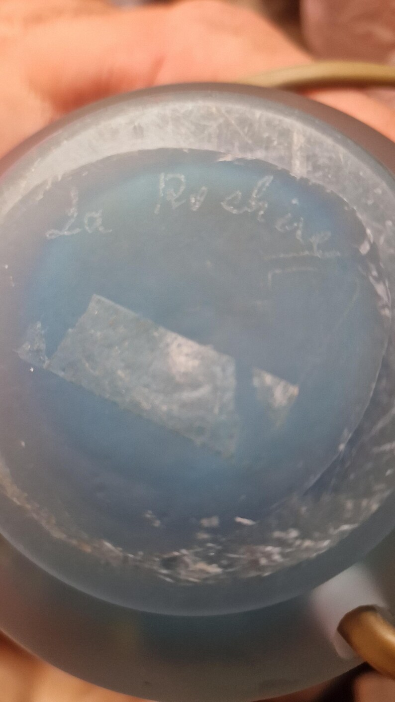 May include: A close-up of a light blue glass object with the handwritten text "La Roch&egrave;re". The object has a slightly textured surface and a small, rectangular piece of tape is visible.