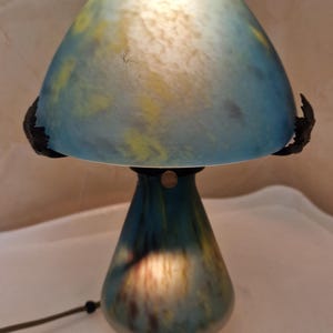 May include: A mushroom-shaped table lamp with a blue and yellow glass shade. The lamp has a dark base and a black cord. The shade is a light blue color with yellow accents. The lamp is lit from within, creating a warm glow.