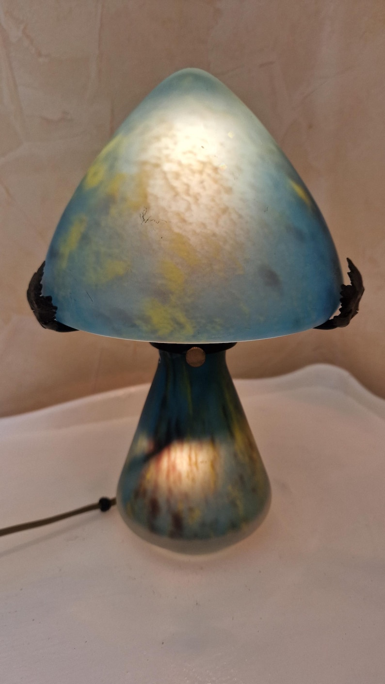 May include: A mushroom-shaped table lamp with a blue and yellow glass shade. The lamp has a dark base and a black cord. The shade is a light blue color with yellow accents. The lamp is lit from within, creating a warm glow.