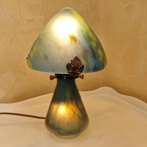 May include: A mushroom-shaped table lamp with a teal and yellow glass shade. The lamp has a dark teal base and a bronze leaf detail. The lamp is lit, casting a warm glow.