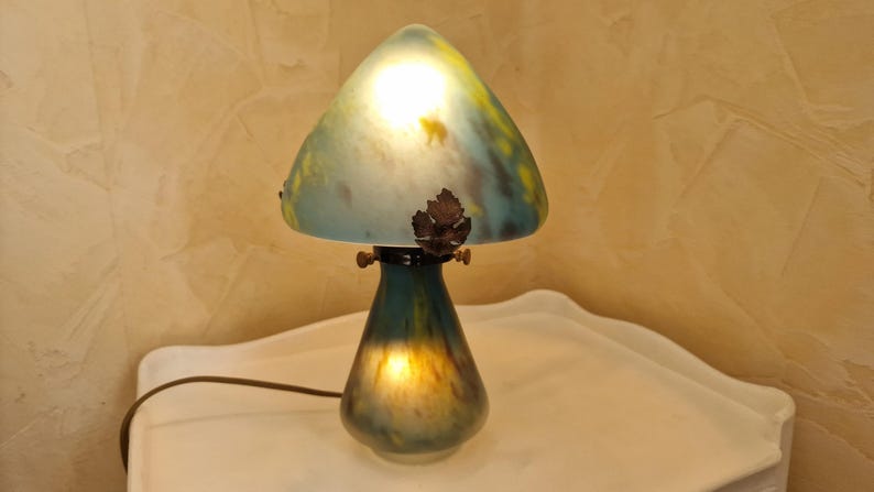 May include: A mushroom-shaped table lamp with a teal and yellow glass shade. The lamp has a dark teal base and a bronze leaf detail. The lamp is lit, casting a warm glow.