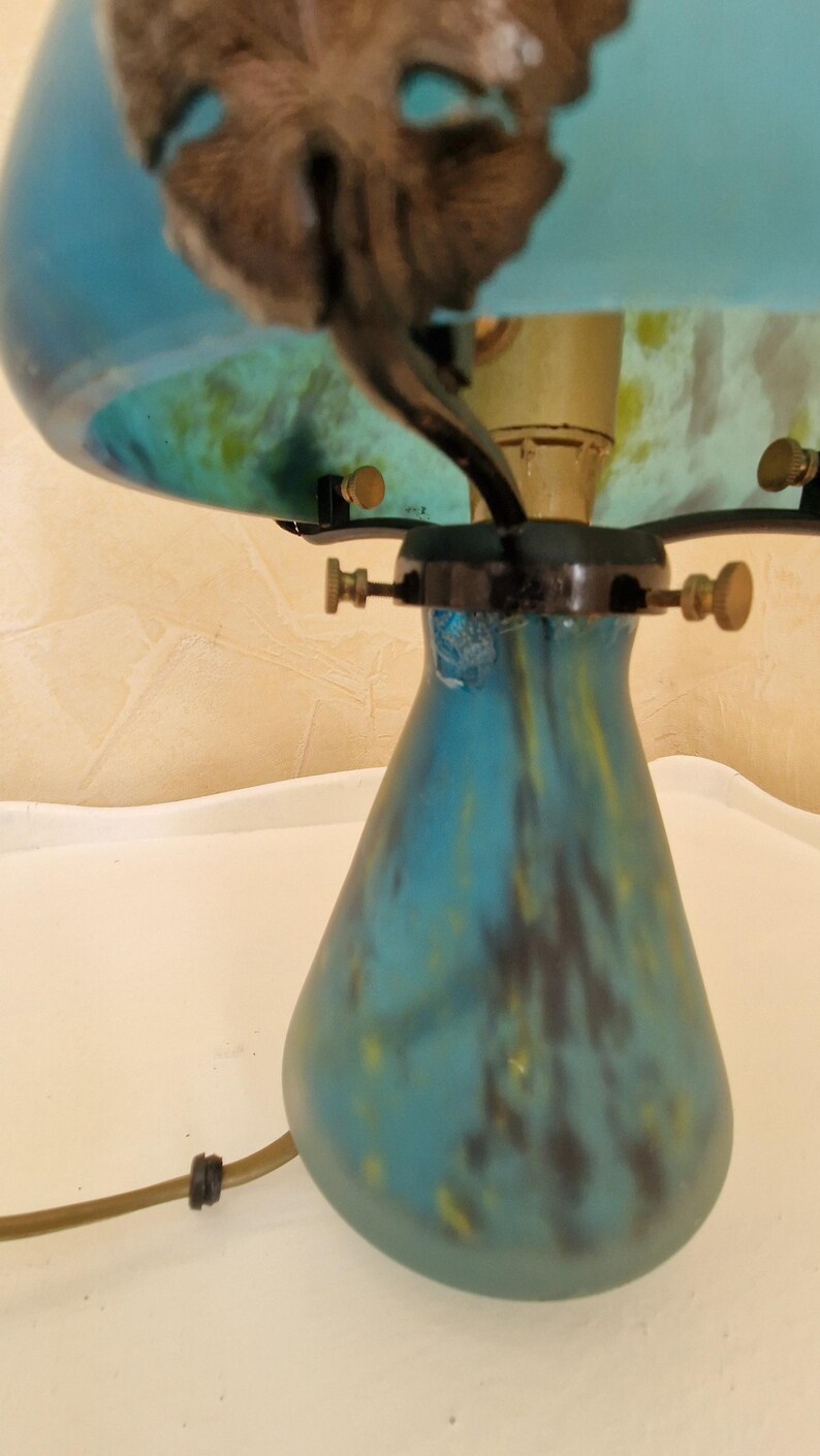 May include: A vintage-style table lamp with a blue and yellow glass shade and base. The lamp features a decorative leaf design on top. The base has a teardrop shape. The lamp has a brass-colored stem and hardware.