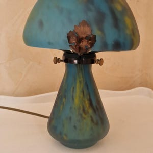 May include: A decorative mushroom-shaped lamp with a teal and yellow glass shade. The lamp has a bronze-colored leaf accent and a dark base. The lamp is on a white surface.