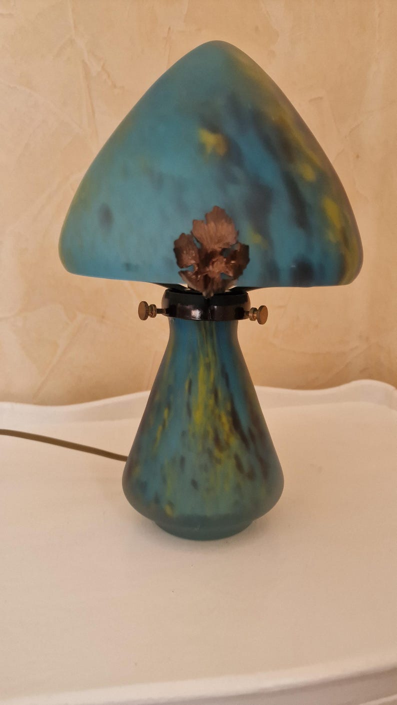 May include: A decorative mushroom-shaped lamp with a teal and yellow glass shade. The lamp has a bronze-colored leaf accent and a dark base. The lamp is on a white surface.