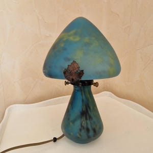 May include: A mushroom-shaped table lamp with a blue and yellow glass shade and a matching base. The lamp has a bronze leaf detail and a brown cord. The lamp is on a white surface.