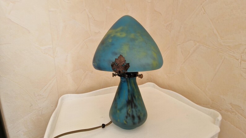 May include: A mushroom-shaped table lamp with a blue and yellow glass shade and a matching base. The lamp has a bronze leaf detail and a brown cord. The lamp is on a white surface.