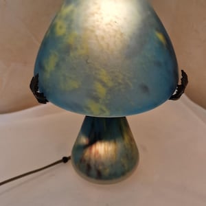 May include: A mushroom-shaped lamp with a blue and yellow glass shade. The lamp has a dark base and a black cord. The shade is lit from within, creating a soft glow. The lamp is on a white surface.