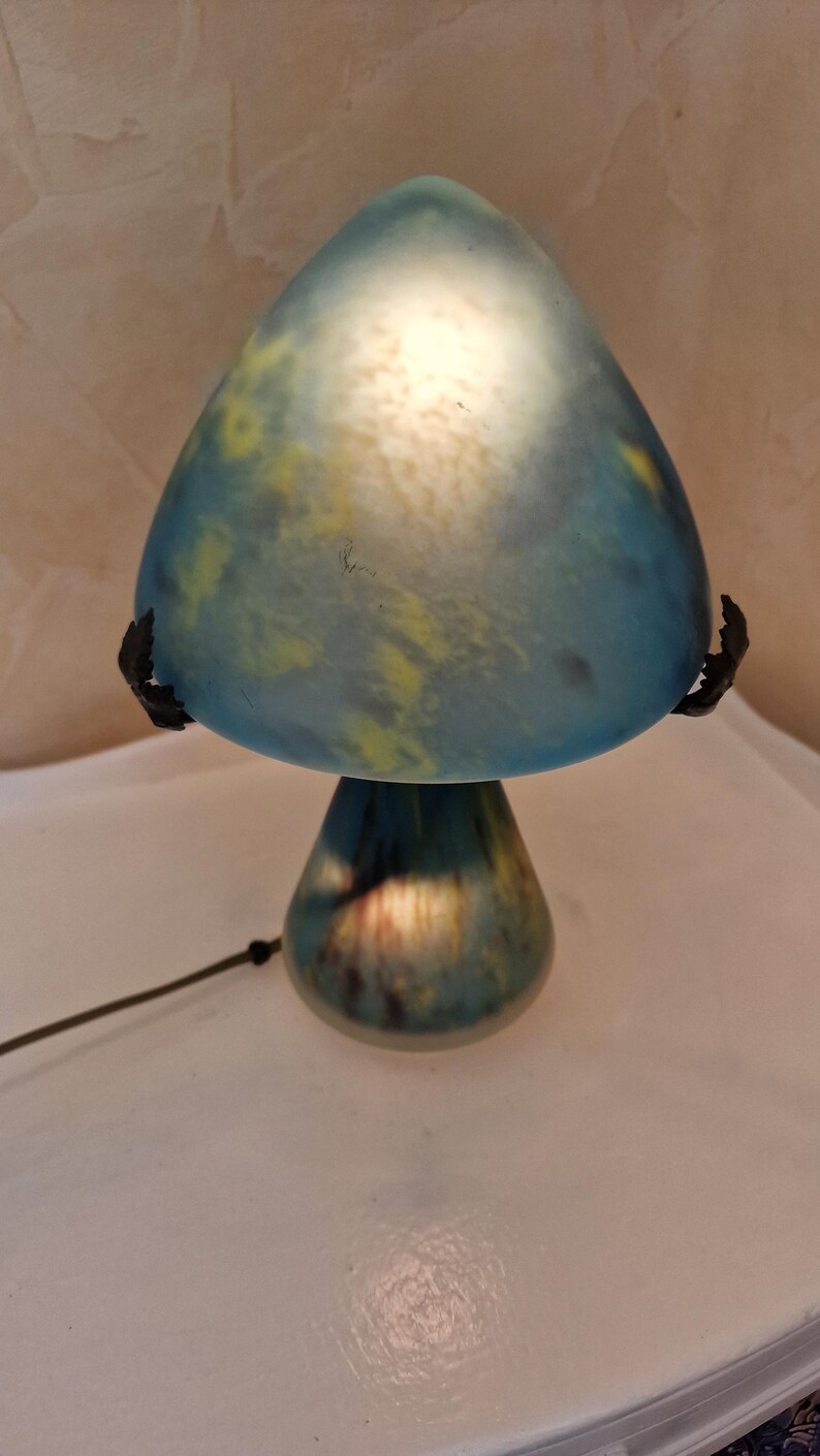 May include: A mushroom-shaped lamp with a blue and yellow glass shade. The lamp has a dark base and a black cord. The shade is lit from within, creating a soft glow. The lamp is on a white surface.