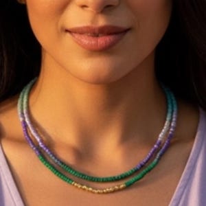 Könnte beinhalten: Nahaufnahme einer Person, die zwei Halsketten übereinander trägt. Die obere Halskette besteht aus einer Abstufung von blauen und grünen Perlen, während die untere Halskette grüne Perlen mit goldenen Akzenten aufweist. Die Person trägt ein lila Oberteil.