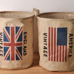 Op de afbeelding: Twee beige stoffen opbergmanden met handvatten. Eén mand toont de Britse vlag met de tekst "Vintage England". De andere toont de Amerikaanse vlag met de tekst "Vintage America". Beide hebben een cilindrische vorm.