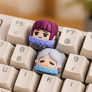 May include: Two custom keycaps with anime-style character designs. One has purple hair, the other white, both with blue scarves. They sit on a white keyboard, with the F5 and F7 keys visible.