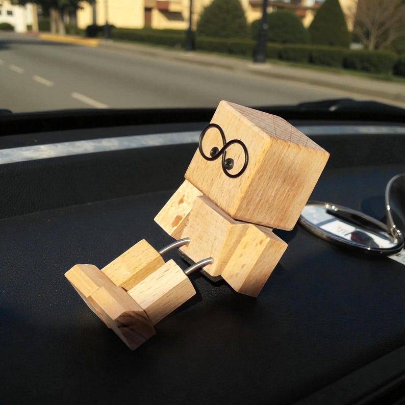 May include: A wooden figure with a block-shaped head and body, and jointed limbs. The figure has black wire-rimmed glasses and is sitting on a black surface. The figure is made of light-colored wood.