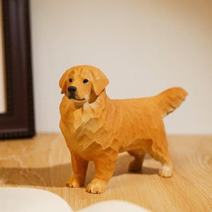 May include: A wooden sculpture of a Golden Retriever dog, carved with visible wood grain. The dog is standing on a wooden surface, with a book and a framed picture in the background. The dog is approximately 8 inches long.