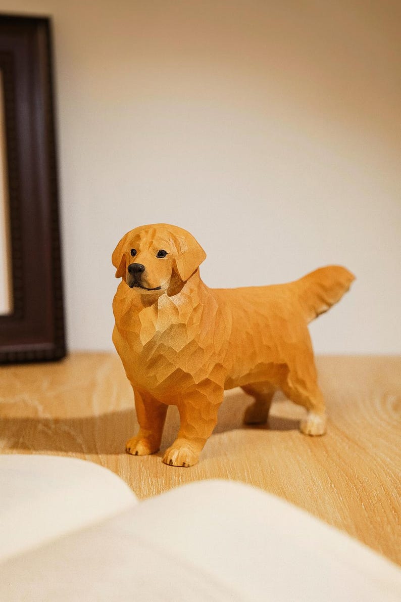 May include: A wooden sculpture of a Golden Retriever dog, carved with visible wood grain. The dog is standing on a wooden surface, with a book and a framed picture in the background. The dog is approximately 8 inches long.