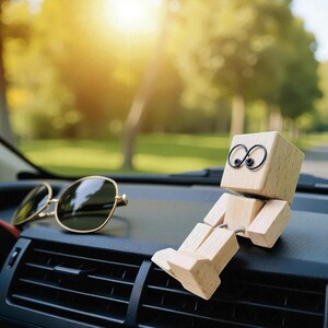 May include: A wooden block figure with wire eyes and jointed limbs sits on a car dashboard next to a pair of gold-rimmed sunglasses. The figure is made of light-colored wood and has a simple, minimalist design.