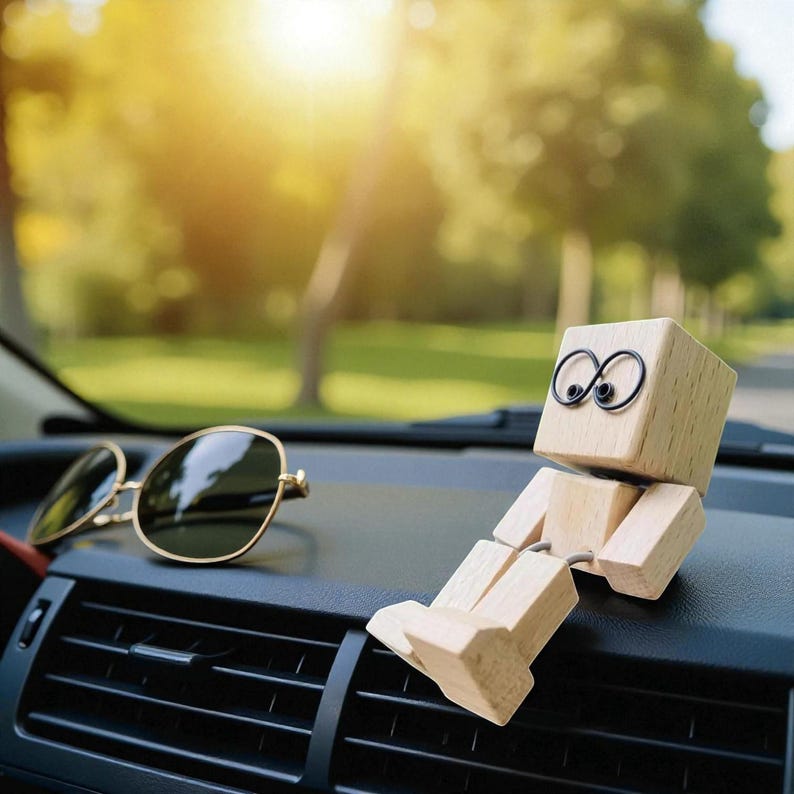 May include: A wooden block figure with wire eyes and jointed limbs sits on a car dashboard next to a pair of gold-rimmed sunglasses. The figure is made of light-colored wood and has a simple, minimalist design.