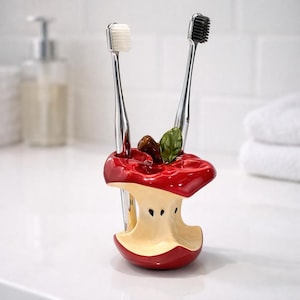 May include: A ceramic toothbrush holder in the shape of a bitten apple, with a red exterior and cream interior. It holds two toothbrushes, one with a white head and one with a black head. The holder is decorated with a leaf and two small brown objects.