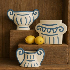 May include: Three off-white ceramic vases with blue detailing and two lemons are displayed on wooden shelves. The vases feature distinctive shapes, including a handled urn and a column design. The blue accents provide a decorative element to the pottery.