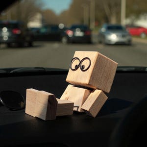 May include: A wooden figure with a cube-shaped head and body, and rectangular limbs. The figure has black wire-framed eyes and is positioned as if reclining. The figure is sitting on a black dashboard, with cars visible in the background.