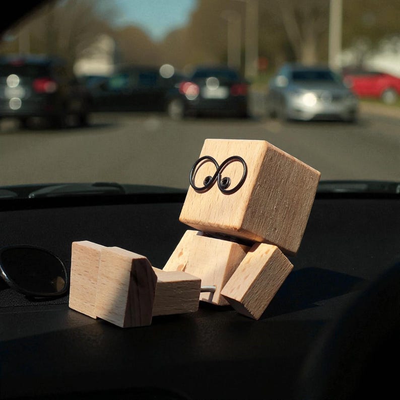 May include: A wooden figure with a cube-shaped head and body, and rectangular limbs. The figure has black wire-framed eyes and is positioned as if reclining. The figure is sitting on a black dashboard, with cars visible in the background.