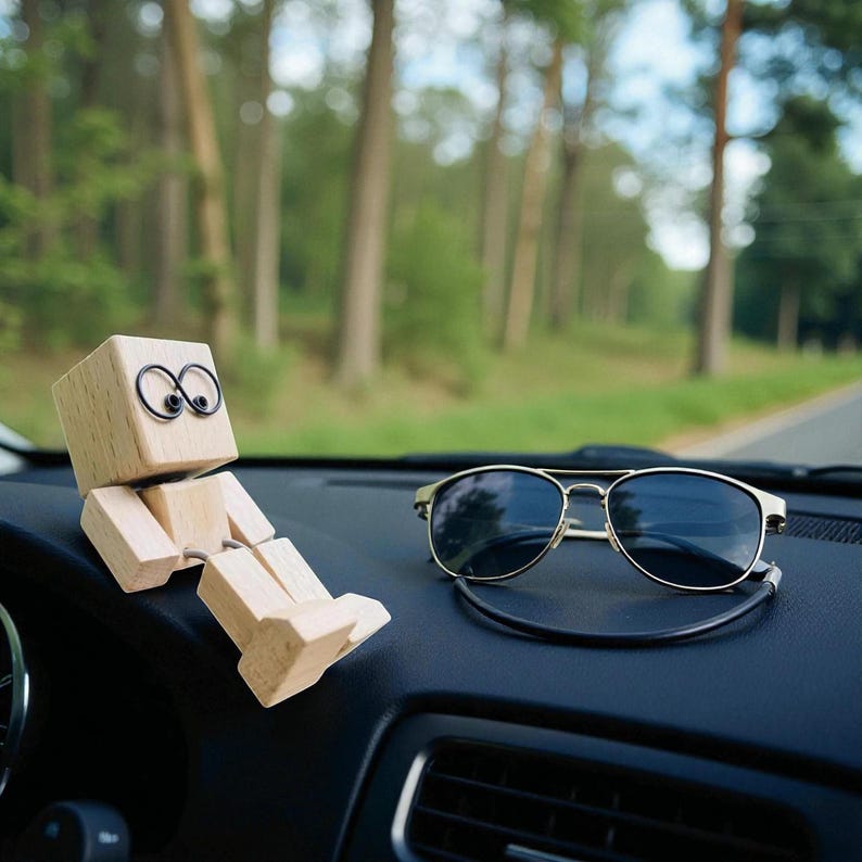 May include: A wooden articulated figure with wire eyes and a pair of gold-framed sunglasses rest on a car dashboard. The figure is made of light-colored wood and is posed in a seated position. The sunglasses have dark lenses.