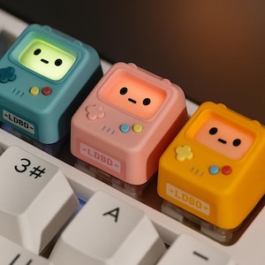 Puede incluir: Tres teclas de teclado coloridas con forma de consolas de juegos portátiles retro. Las teclas son de color turquesa, rosa y amarillo, cada una con una pequeña pantalla que muestra una cara sonriente. También tienen botones y el texto "-LDBO-".