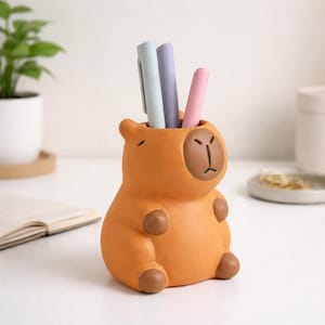 Op de afbeelding: Een oranje capibara-vormige pennenhouder met drie pennen. De pennenhouder heeft een lichtbruin gezicht en poten. De pennen zijn lichtblauw, lila en roze.