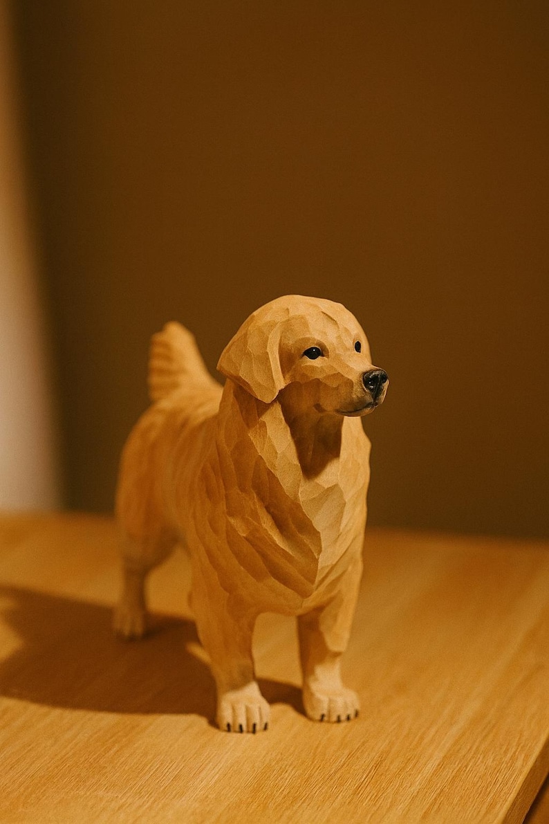 May include: A wooden sculpture of a Golden Retriever dog, standing on all fours. The dog is carved from light-colored wood, with detailed features including its fur, eyes, and nose. The sculpture is displayed on a wooden surface.
