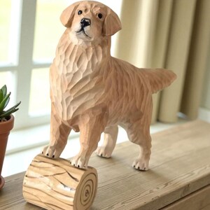 May include: A carved wooden sculpture of a Golden Retriever dog standing on a log. The dog is light brown with detailed carving, and its front paws rest on a small, round log. The sculpture is displayed on a wooden surface.