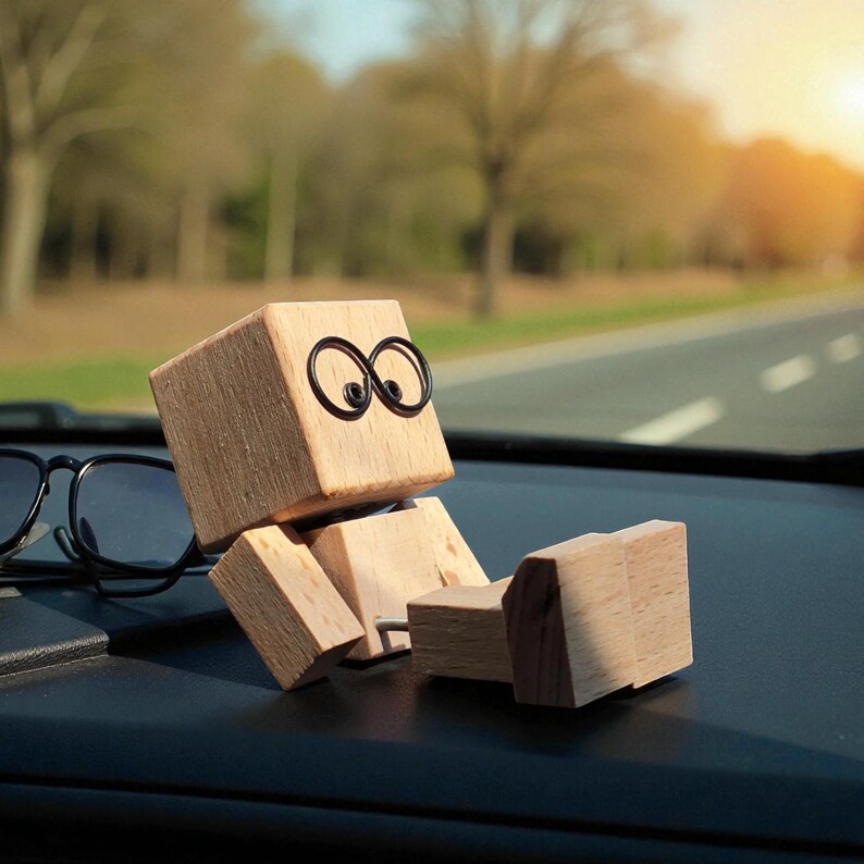 May include: A wooden figure with a cube-shaped head and jointed body, featuring black wire-framed eyes. The figure is positioned on a car dashboard, with a road and trees visible in the background. The figure is made of light-colored wood.