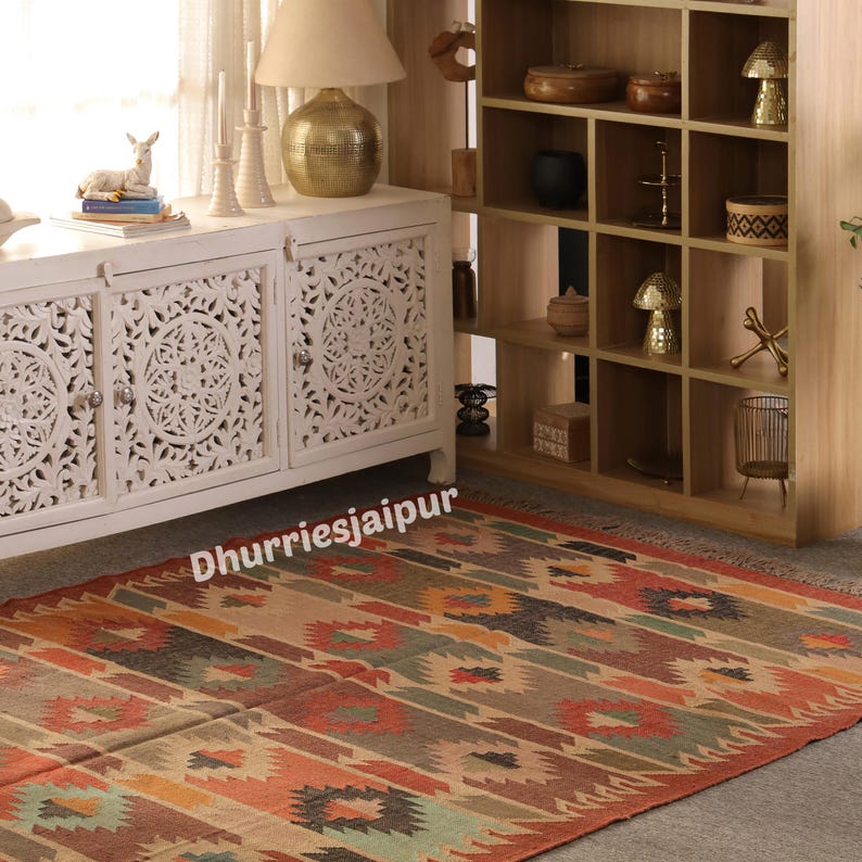 May include: A colorful, patterned rug with a geometric design in shades of red, orange, green, and brown. The rug is placed on a neutral-colored floor, with a white cabinet and wooden shelving unit in the background. The text "Dhurriesjaipur" is visible on the rug.