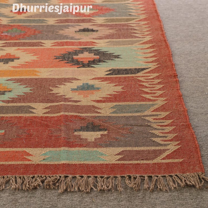 May include: A rectangular rug with a geometric pattern in shades of red, orange, brown, and teal. The rug has a fringed edge and the text "Dhurriesjaipur" in the upper left corner. The rug is made of a woven material.
