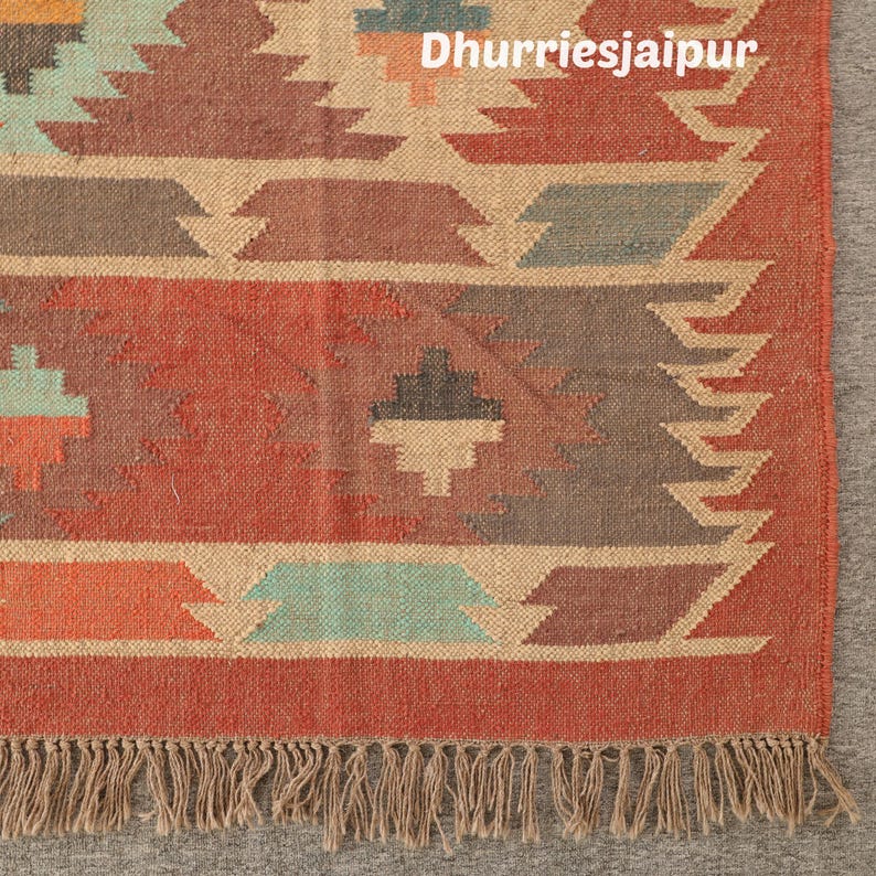 May include: Close-up of a handwoven rug with a geometric pattern in shades of red, orange, beige, and teal. The rug has a fringed edge and the text "Dhurriesjaipur" is visible.