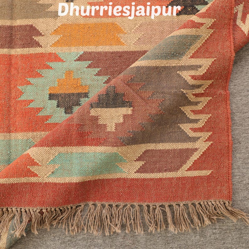 May include: A colorful woven rug with a geometric pattern in shades of red, orange, teal, and beige. The rug has a fringed edge and the text "Dhurriesjaipur" is visible at the top. The rug is partially rolled up, revealing the underside.