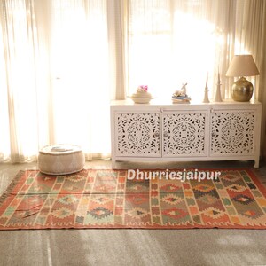 May include: A rectangular rug with a geometric pattern in shades of orange, beige, and teal. The rug is placed on a gray floor in front of a white cabinet with carved doors. The text "Dhurriesjaipur" is visible on the rug.
