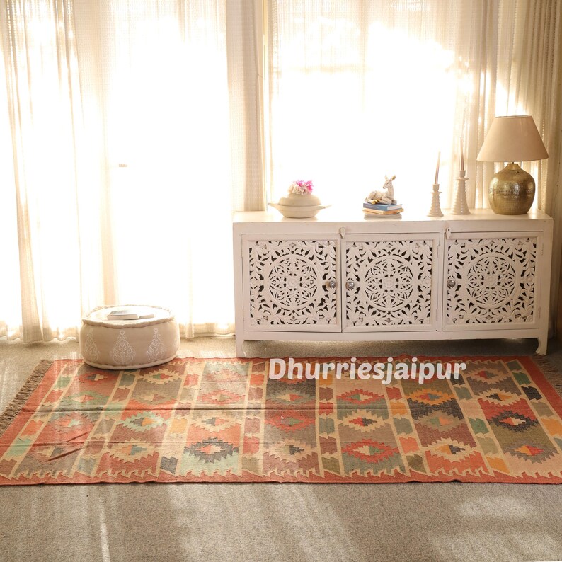 May include: A rectangular rug with a geometric pattern in shades of orange, beige, and teal. The rug is placed on a gray floor in front of a white cabinet with carved doors. The text "Dhurriesjaipur" is visible on the rug.