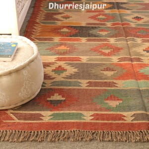 May include: A colorful, rectangular rug with a geometric pattern in shades of red, brown, green, and beige. The rug has a fringed edge and the text "Dhurriesjaipur" is visible at the top. The rug is placed on a gray surface.