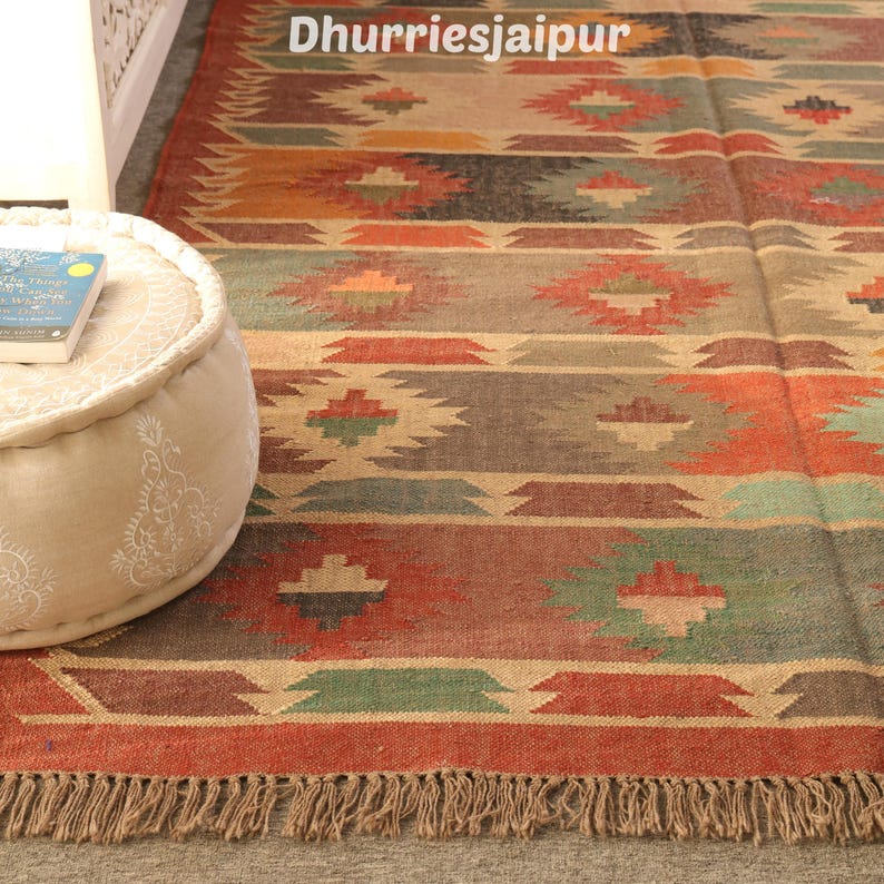 May include: A colorful, rectangular rug with a geometric pattern in shades of red, brown, green, and beige. The rug has a fringed edge and the text "Dhurriesjaipur" is visible at the top. The rug is placed on a gray surface.
