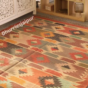 May include: A colorful, patterned rug with a geometric design. The rug features a mix of warm colors, including orange, red, brown, and green, arranged in horizontal stripes and floral-like motifs. The text "Dhurriesjaipur" is visible on the rug.