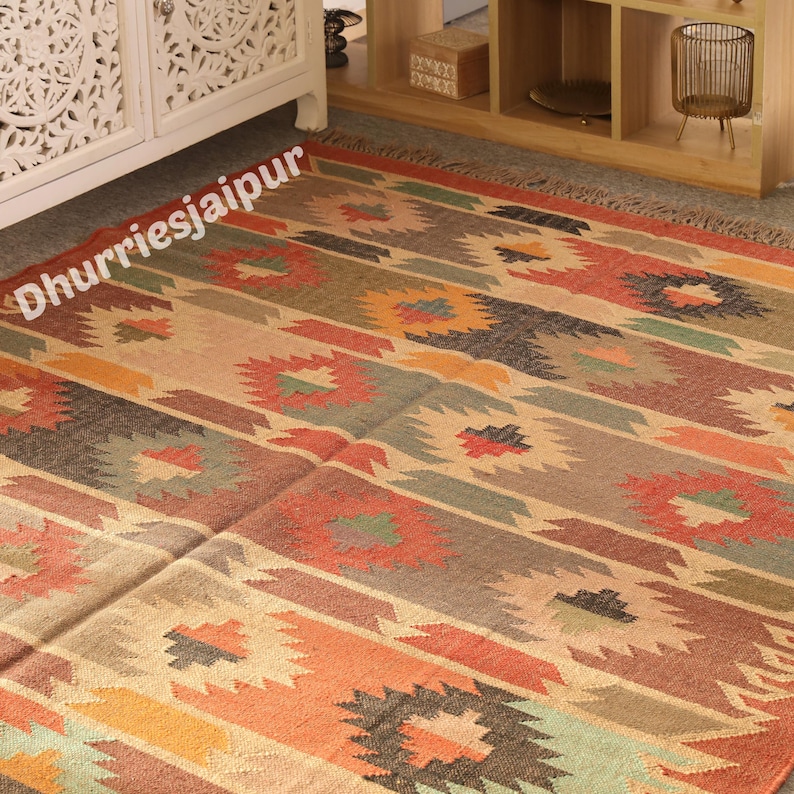 May include: A colorful, patterned rug with a geometric design. The rug features a mix of warm colors, including orange, red, brown, and green, arranged in horizontal stripes and floral-like motifs. The text "Dhurriesjaipur" is visible on the rug.