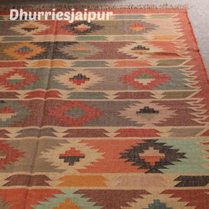 May include: A colorful Dhurrie rug with a geometric pattern in shades of red, orange, green, and beige. The rug features a repeating diamond and stripe design, with the text "Dhurriesjaipur" in the upper left corner.