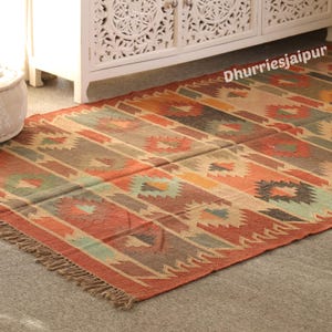 May include: A rectangular rug with a geometric pattern in shades of red, orange, brown, and beige. The rug has a fringed edge and the text "Dhurriesjaipur" is visible in the upper right corner. The rug is placed on a gray surface.